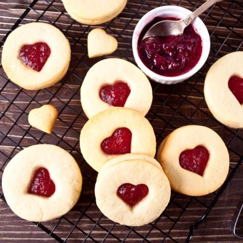 Sablés à la confiture de fraise sur un plan de travail en bois, centre des sablés en forme de coeur, saupoudrés de sucre glace, ramequin blanc avec de la confiture de fraise et une cuillere à café dedans