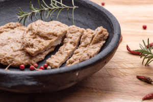 Seitan maison, fausse viande végane, présentée sous forme d'escalopes dans une assiette creuse noire avec une branche de romarin et des baies