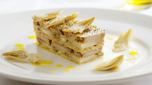 Feuilleté de foie gras et artichaut, avec des couches de coeur d'artichaut et des tranches fines de foie gras en alternance, servi sur une assiette blanche moderne