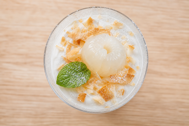Verrine de mousse litchi coco avec un litchi, noix de coco râpée et menthe, sur fond bois clair.